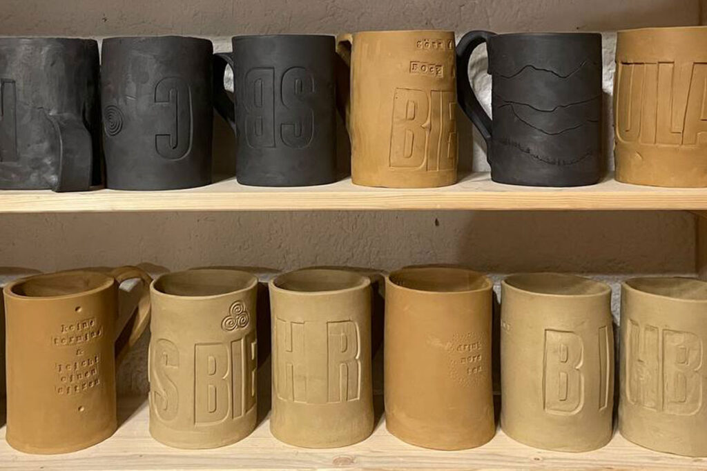 Handgefertigte Bierkrüge in Schwarz und Naturton auf Holzregalen, mit eingeprägten Buchstaben und grafischen Strukturen, ausgestellt im Atelier