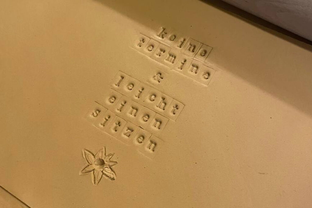 Detailaufnahme einer Tonfläche mit eingeprägtem Schriftzug ‚keine termine & leicht einen sitzen‘ und kleiner Blume darunter.