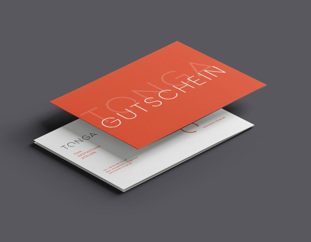 Minimalistischer Geschenkgutschein des Atelier Tonga: eine rote Karte mit dem weißen Schriftzug ‚GUTSCHEIN‘ und dem dezenten Hintergrundtext ‚TONGA‘ liegt auf einer hellen Karte mit Kontaktdaten des Ateliers. Elegantes, modernes Design auf dunklem Untergrund.